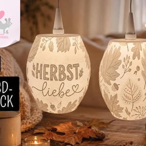 Könnte beinhalten: Zwei weiße Hängelampenschirme mit einem 3D-gedruckten Design aus Herbstblättern und Eicheln. Ein Schirm zeigt den Text "HERBST liebe" mit einem Herzsymbol. Die Lampenschirme sind zylindrisch mit einer verjüngten Oberseite und an weißen Kabeln aufgehängt.