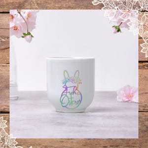Może przedstawiać: Biały kubek ceramiczny z tęczowym kr&oacute;likiem i tekstem "Frohe Ostern" w tęczowych kolorach.