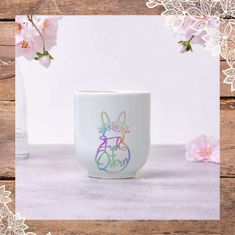Może przedstawiać: Biały kubek ceramiczny z tęczowym kr&oacute;likiem i tekstem "Frohe Ostern" w tęczowych kolorach.