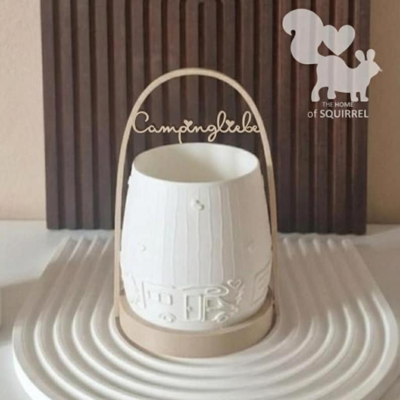 Op de afbeelding: Een witte keramische lantaarn met een houten handvat en voet. De lantaarn heeft een textuur met reli&euml;f details van huizen en harten. De tekst "Campingliebe" staat op het handvat. Een eekhoornlogo is zichtbaar op de achtergrond.