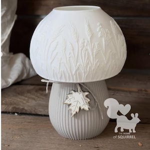 May include: A white mushroom-shaped lamp with a dome top featuring embossed wheat stalks. The base is light grey with vertical ridges and is tied with a thin string holding a white maple leaf charm. The text THE HOME of SQUIRREL is visible.