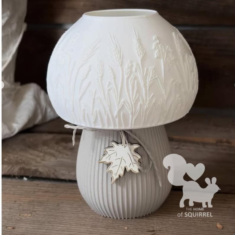 May include: A white mushroom-shaped lamp with a dome top featuring embossed wheat stalks. The base is light grey with vertical ridges and is tied with a thin string holding a white maple leaf charm. The text THE HOME of SQUIRREL is visible.