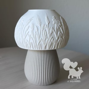 May include: A white mushroom-shaped lamp with a textured dome featuring embossed wheat stalks and grasses. The base is light grey with vertical ribbed detailing. A logo with a squirrel silhouette and the text THE HOME of SQUIRREL is visible.