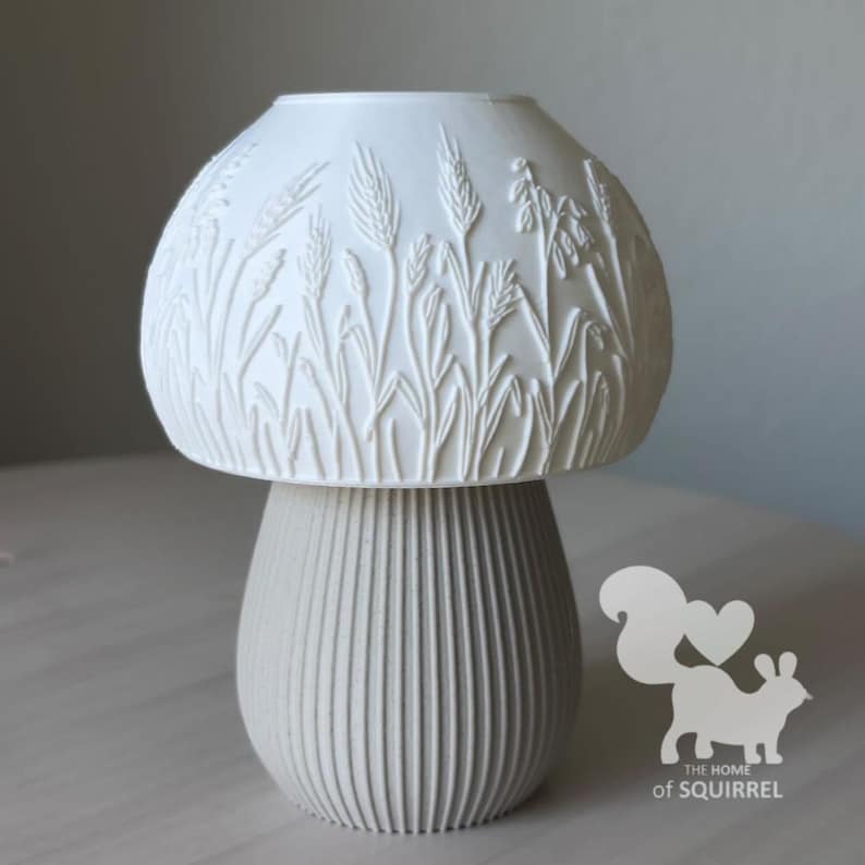 May include: A white mushroom-shaped lamp with a textured dome featuring embossed wheat stalks and grasses. The base is light grey with vertical ribbed detailing. A logo with a squirrel silhouette and the text THE HOME of SQUIRREL is visible.