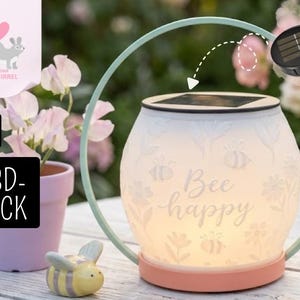 Puede incluir: Una linterna solar con un cuerpo de vidrio blanco con estampado floral y una base color melocotón. La linterna tiene un asa verde menta y las palabras "Bee happy" están escritas en la parte delantera. Una pequeña figura de abeja de cerámica está en primer plano.