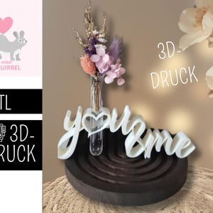 May include: A white 3D-printed "you & me" decorative piece with a heart-shaped accent, holding a small glass vase with dried flowers. The piece sits on a dark brown, semi-circular base. The image also includes text that says ".STL" and "3D-DRUCK".