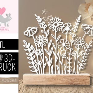May include: A white, laser-cut floral arrangement featuring daisies and other wildflowers, mounted on a rectangular wooden base. The design includes delicate leaves and stems. The words "3D-DRUCK" are visible.