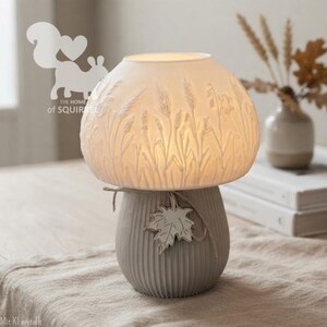 May include: A mushroom-shaped table lamp with a white translucent shade featuring embossed wheat stalks. The base is a ribbed grey ceramic cylinder decorated with a small white maple leaf charm tied on with a thin piece of twine string.
