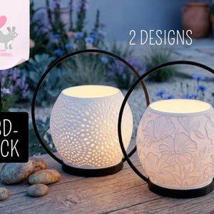 May include: Two white ceramic lanterns with black metal handles. One lantern has a perforated design, the other features a floral pattern. The lanterns are lit from within, creating a warm glow. The text "2 DESIGNS" is visible.