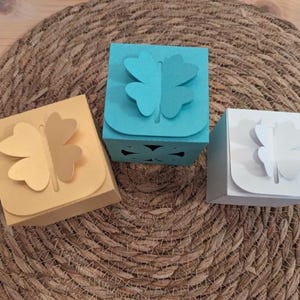 Pu&ograve; includere: Tre piccole scatole regalo quadrate nei toni del giallo, verde acqua e bianco, ognuna adornata con un motivo a quadrifoglio ritagliato sul coperchio. Le scatole sono disposte su un sottobicchiere rotondo e intrecciato.
