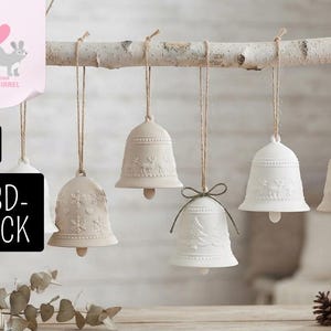 Può includere: Una collezione di sei ornamenti a forma di campana color crema e bianco sporco, appesi a un ramo di betulla tramite spago. Le campane presentano motivi in rilievo di renne, fiocchi di neve e pini. Una campana è legata con un nastro verde.