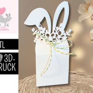 Könnte beinhalten: Eine weiße Osterhasen-Geschenkbox aus Papier mit dekorativen floralen Ausschnitten und einer grün-weißen Schnur-Schleife. Die Box steht auf einer braunen Oberfläche. Das Bild enthält auch ein Logo mit einem Eichhörnchen und dem Text ".STL 3D-DRUCK".