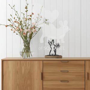 Op de afbeelding: Een zwartmetalen konijnenbeeldje met een bloem, op een houten voet. Het konijn staat voor een witte muur met een konijnensilhouet. Een glazen vaas met roze bloemen staat links op een houten kast.