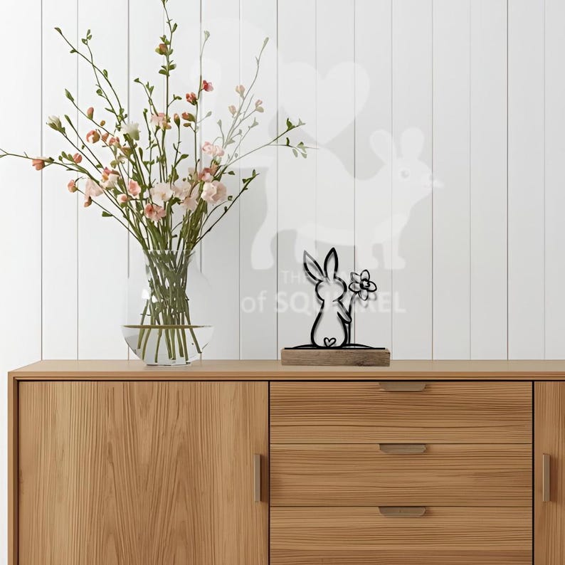 Op de afbeelding: Een zwartmetalen konijnenbeeldje met een bloem, op een houten voet. Het konijn staat voor een witte muur met een konijnensilhouet. Een glazen vaas met roze bloemen staat links op een houten kast.