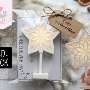 May include: A white star-shaped ornament with an intricate cutout floral pattern, mounted on a small stand. The ornament is packaged in a clear plastic bag with a cardboard backing labeled WEIHNACHTSDEKO to go and a brown tag reading Frohe Weihnachten.