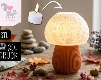 Könnte beinhalten: Eine pilzförmige Lampe mit einem gerippten orangefarbenen Sockel und einem halbtransparenten weißen Schirm, der mit eingravierten Blättern, Eicheln und einem Igel verziert ist, sowie dem Schriftzug "HERBST licht", beleuchtet durch ein weißes Teelicht.