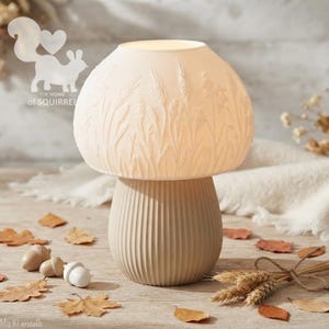 May include: A mushroom-shaped table lamp with a cream-coloured, ribbed base and a translucent, off-white dome shade featuring an embossed pattern of wheat stalks and grasses. The lamp emits a soft, warm glow from within the shade.