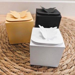 Pu&ograve; includere: Tre scatole regalo quadrate in giallo, nero e bianco, ognuna adornata con una decorazione a forma di farfalla sul coperchio. Le scatole sono disposte su un tappetino circolare intrecciato, creando una presentazione affascinante.