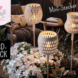 May include: Three white, decorative candle holders with a perforated design, set in a garden bed with white flowers and succulents. The candle holders are on wooden stakes. The image also includes the text "Mini-Stecker" and a logo with a squirrel.