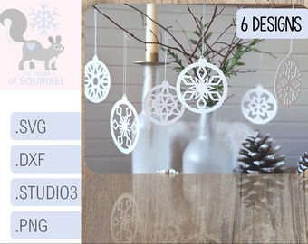 May include: Six white paper snowflake ornaments hanging from a branch. The ornaments are cut out in a variety of snowflake designs. The text "6 DESIGNS" is visible in the upper right corner of the image.