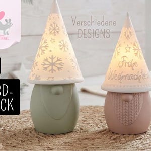 May include: Two gnome-shaped lamps with conical hats. The left hat features a snowflake pattern, while the right hat displays the text "Frohe Weihnachten". The gnomes have sage green and dusty pink bodies, respectively, and are illuminated by tea lights.
