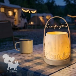 Op de afbeelding: Een verlichte keramische lantaarn met het woord "Campingliebe" en kampeerthema's, waaronder een camper, bomen en harten. Een beige emaille mok staat in de buurt op een blauw-wit geruite tafelkleed. Het logo van The Home of Squirrel is zichtbaar.