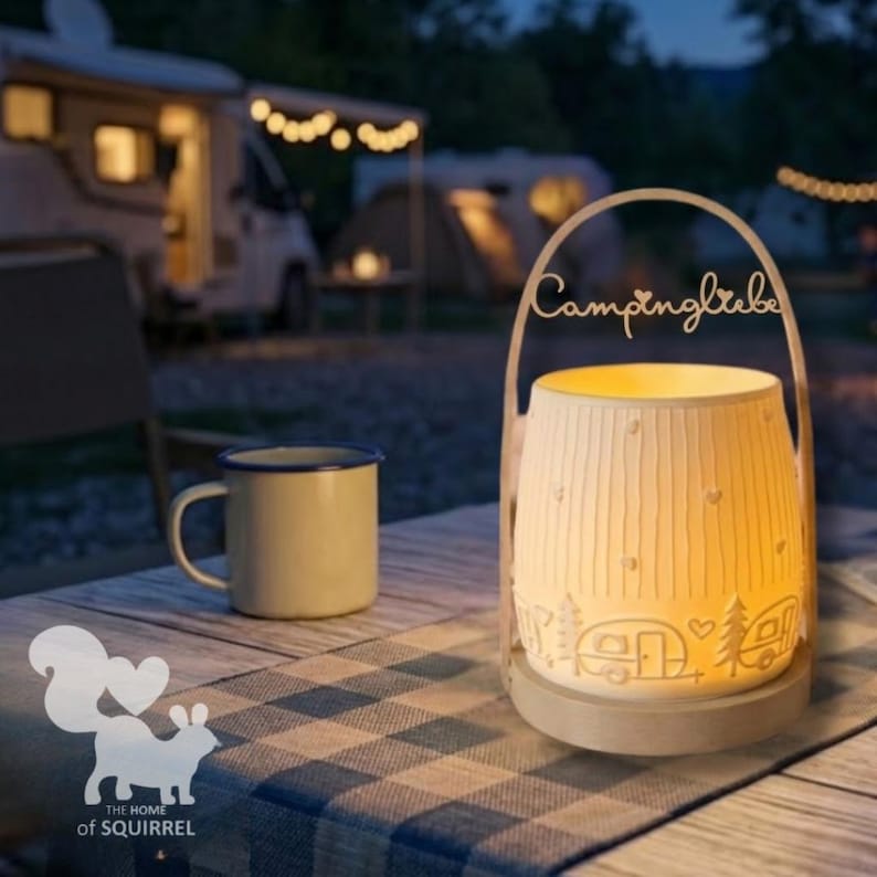 Op de afbeelding: Een verlichte keramische lantaarn met het woord "Campingliebe" en kampeerthema's, waaronder een camper, bomen en harten. Een beige emaille mok staat in de buurt op een blauw-wit geruite tafelkleed. Het logo van The Home of Squirrel is zichtbaar.