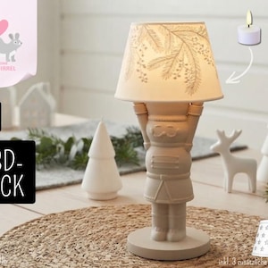 May include: A beige nutcracker-shaped table lamp with a conical lampshade featuring a pine branch pattern. The lamp is designed for a tea light candle. Three additional interchangeable lampshades with star and geometric patterns are included.