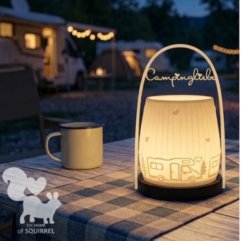 Op de afbeelding: Een witte lantaarn met de tekst "Campingliebe" en een kampeerthema, inclusief een camper, bomen en harten. Een gele mok met een blauwe rand staat op een geruite tafelkleed. De achtergrond toont een camping in de schemering.