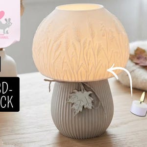 Könnte beinhalten: Eine pilzförmige Lampe mit einem weißen, durchscheinenden Schirm mit geprägten Weizenähren und einem gerippten, hellgrauen Fuß, der mit einem Ahornblatt verziert ist. Rechts neben der Lampe steht ein kleines weißes Teelicht.