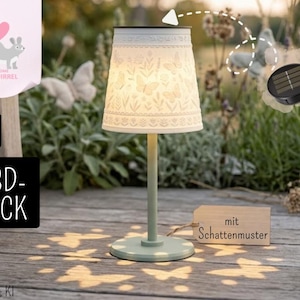 Op de afbeelding: Een op zonne-energie werkende tafellamp met een witte lampenkap met een vlinder- en bloemenpatroon. De lamp werpt vlindervormige schaduwen. De voet en steel zijn lichtgroen. De tekst "mit Schattenmuster" is ook zichtbaar.