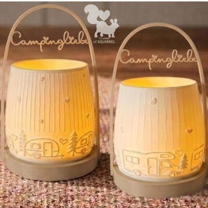 Op de afbeelding: Twee cr&egrave;mekleurige keramische lantaarns met een campingthema. Elke lantaarn heeft een cilindrische vorm met verticale groeven en een decoratieve basis. De lantaarns hebben uitgesneden ontwerpen van campers, bomen en harten. Het woord "Campingliebe" staat in cursief bovenaan.