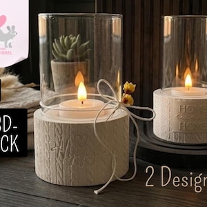 Può includere: Due portacandele bianchi con cilindri di vetro. Uno ha un motivo a trecce e un fiocco, l'altro ha le parole "HOME SWEET HOME". Entrambi contengono una candela accesa. L'immagine include anche una piccola pianta grassa in vaso.