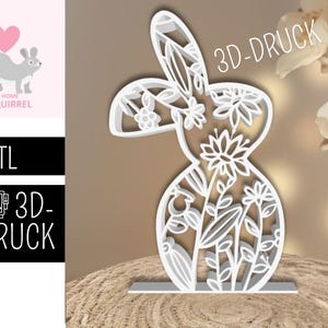 Może przedstawiać: Biała, wycinana laserowo sylwetka królika z kwiatowymi detalami. Królik stoi na małej podstawie. Obraz zawiera również tekst "3D-DRUCK" i logo "THE HOME of SQUIRREL".