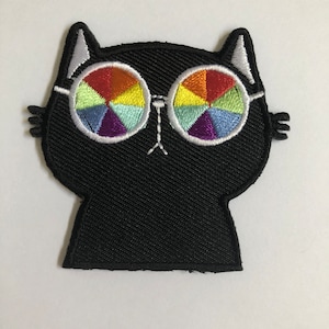 May include: Black cat patch with rainbow-colored glasses. The cat's glasses have a segmented design with colors including red, orange, yellow, green, blue, and purple. The patch is embroidered with white thread outlining the cat and glasses.