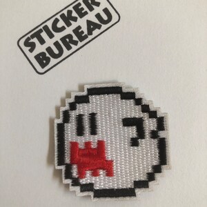 May include: A white and black pixelated embroidered patch of a ghost with a red mouth. The patch is on a white background with a black stamp that says "Sticker Bureau".
