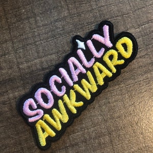 May include: A black embroidered patch with the text "Socially Awkward" in pink and yellow lettering.