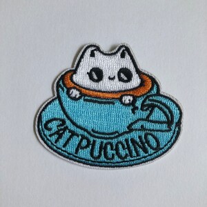 Op de afbeelding: Een blauwe en witte geborduurde patch met een witte kat die in een blauwe koffiekop zit. De tekst "Cat-puccino" is in zwart rond de kop geborduurd.