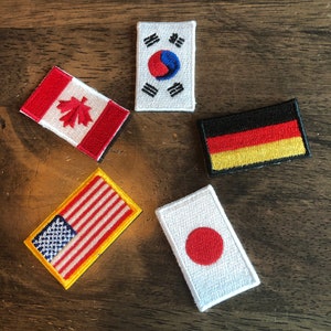 Könnte beinhalten: Fünf gestickte Aufnäher mit den Flaggen der Vereinigten Staaten, Kanadas, Südkoreas, Deutschlands und Japans. Die Aufnäher befinden sich auf einer Holzoberfläche.