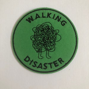以下が含まれることがあります： 黒い刺繍が施された緑色の円形のワッペン。ワッペンの縁には「WALKING DISASTER」という文字が刺繍されている。ワッペンの中央には、もつれた線で体が表現された人の黒い線画がある。