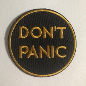 Könnte beinhalten: Ein runder schwarzer Patch mit einem goldenen gestickten Rand. Der Text "DON'T PANIC" ist in Gold gestickt.