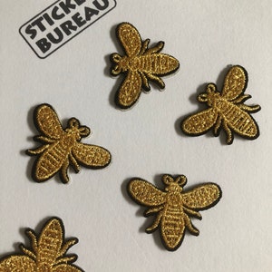 May include: Five gold embroidered bee patches with black outlines. The bees are facing different directions.