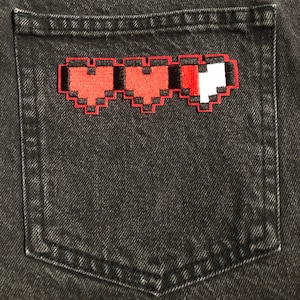 May include: A red and black pixelated heart patch on a black denim pocket.