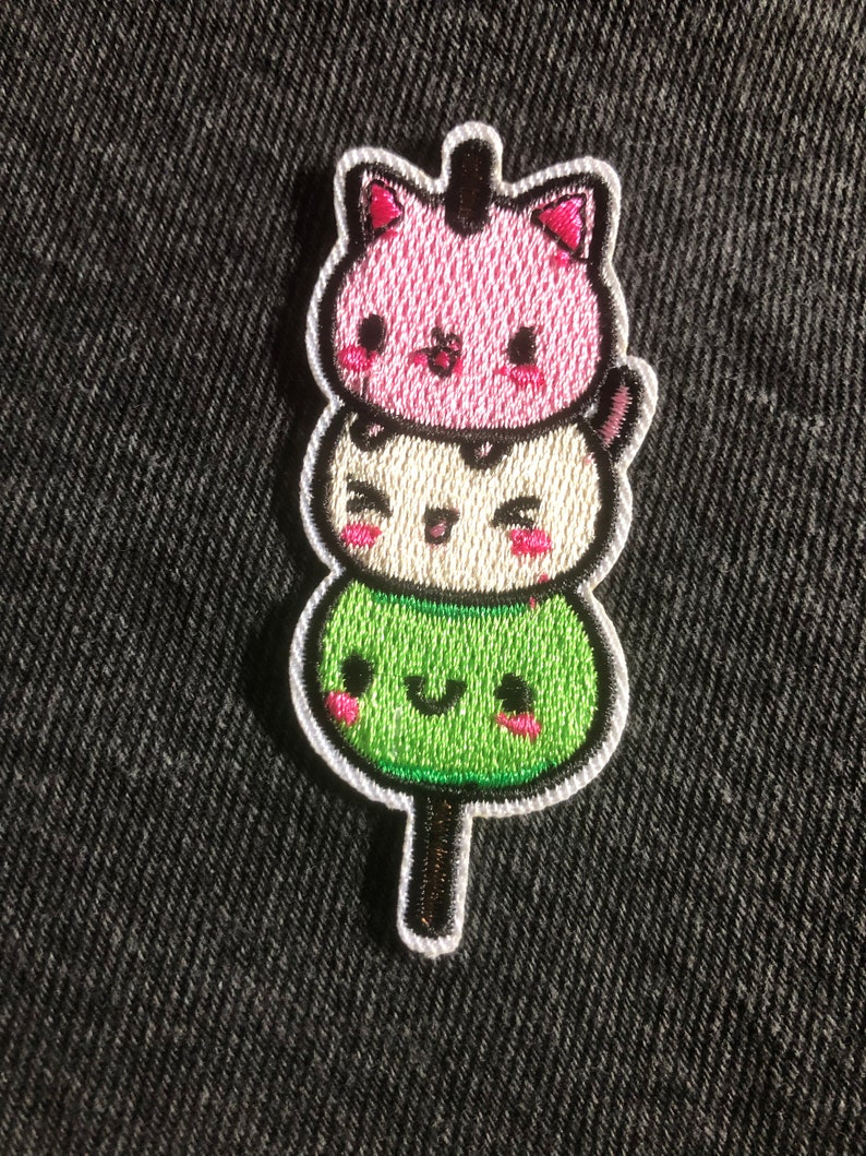 May include: An embroidered patch featuring three stacked kawaii cat faces on a stick. The cat faces are green, white, and pink with smiling expressions and pink cheeks.