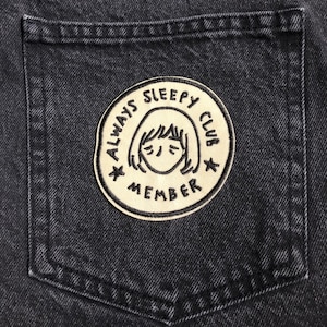 May include: A beige embroidered patch with a cartoon illustration of a girl with her eyes closed. The patch says "Always Sleepy Club Member" in a circle around the girl.