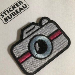 Könnte beinhalten: Gestickter Aufnäher in Form einer Kamera mit hellgrauem Körper, schwarzem Rand und rosa Streifen. Das Objektiv ist hellblau mit schwarzem Zentrum. Ein kleiner rechteckiger Aufkleber mit dem Text "STICKER BUREAU" befindet sich im Hintergrund.