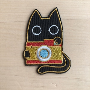 May include: Embroidered patch of a black cat holding a vintage camera. The camera is gold and red with a blue lens. The patch has a gold border and white eye details. The cat's tail is curved.