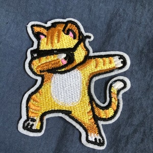 May include: An embroidered iron-on patch of a cartoon cat wearing sunglasses and doing a dabbing pose, in yellow and orange.