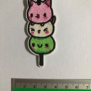 May include: An embroidered patch featuring three stacked kawaii characters on a stick. The top character is a pink cat with a black nose and eyes. The middle character is white with a black nose and eyes. The bottom character is green with a black nose and eyes. The patch is approximately 5 cm tall.