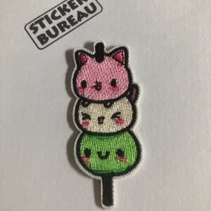 May include: A cute embroidered patch featuring a stack of three green, white, and pink mochi with faces, on a stick.